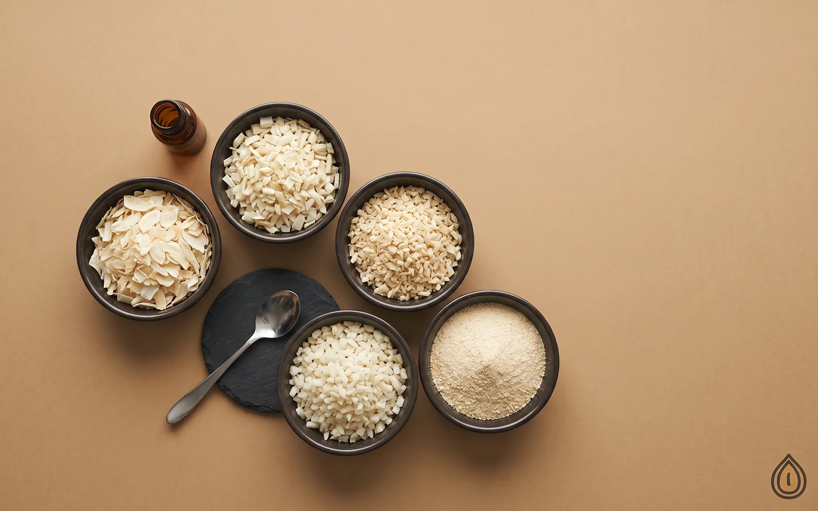 Five dark bowls of dehydrated white onion in decreasing particle size, from large flakes to fine powder, on kraft paper