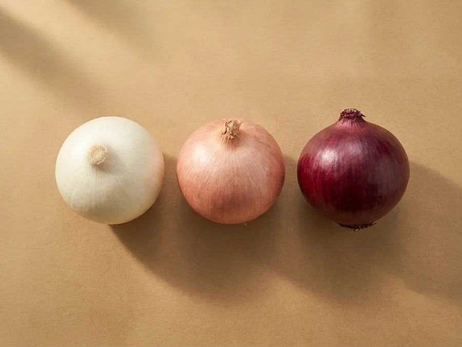 Three whole fresh onions side by side: white, pink and deep red-purple, dry papery skins on kraft paper.