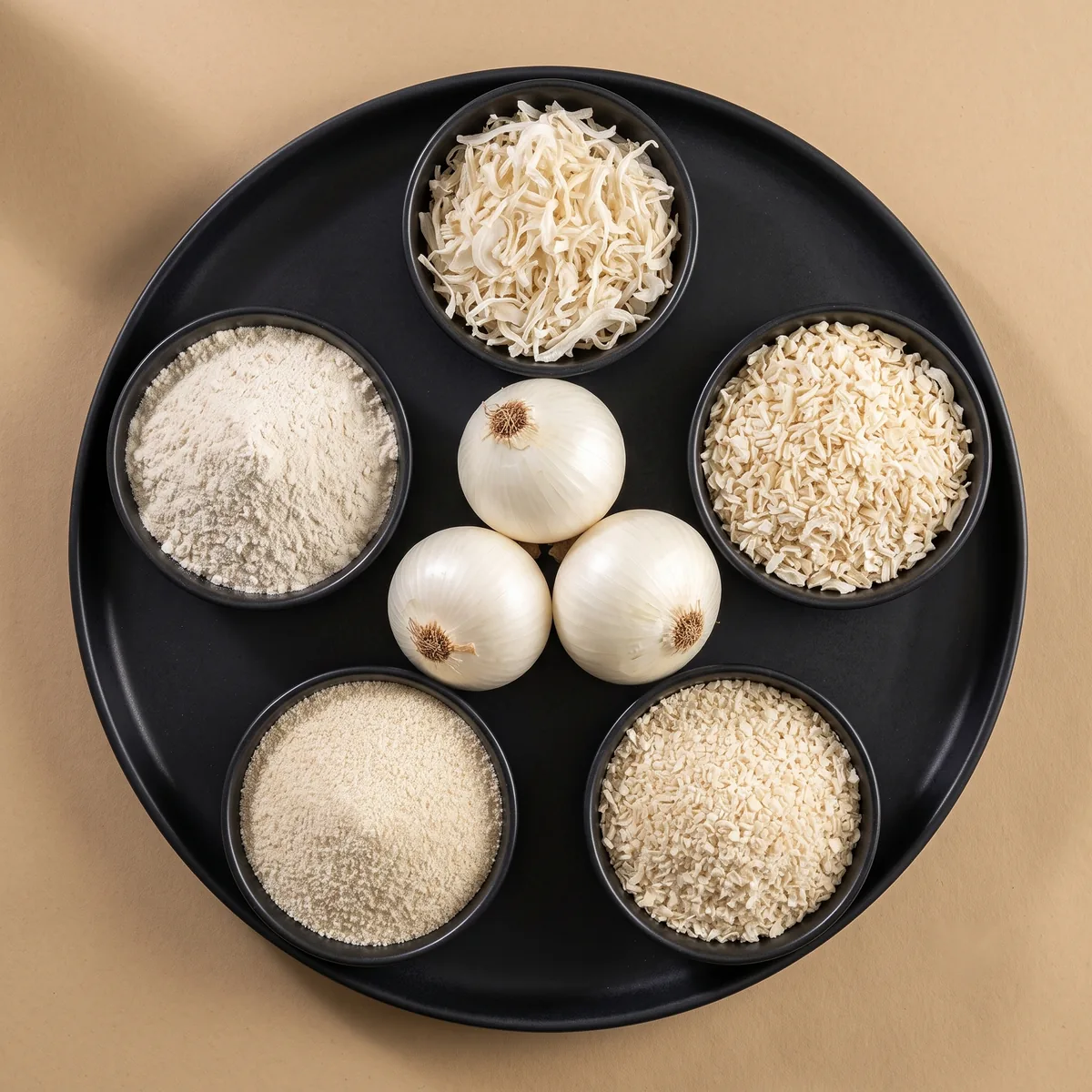 Overhead view of a charcoal platter holding five bowls of dehydrated white onion - flakes, chopped, minced, granules and powder - beside three whole fresh white onions.