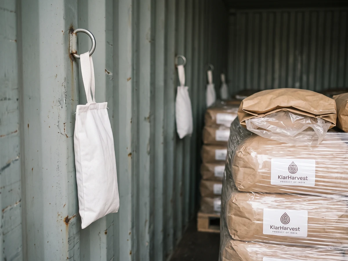 A white desiccant pouch hanging from a lashing ring on a container wall beside a wrapped pallet of kraft sacks with the KlarHarvest label.