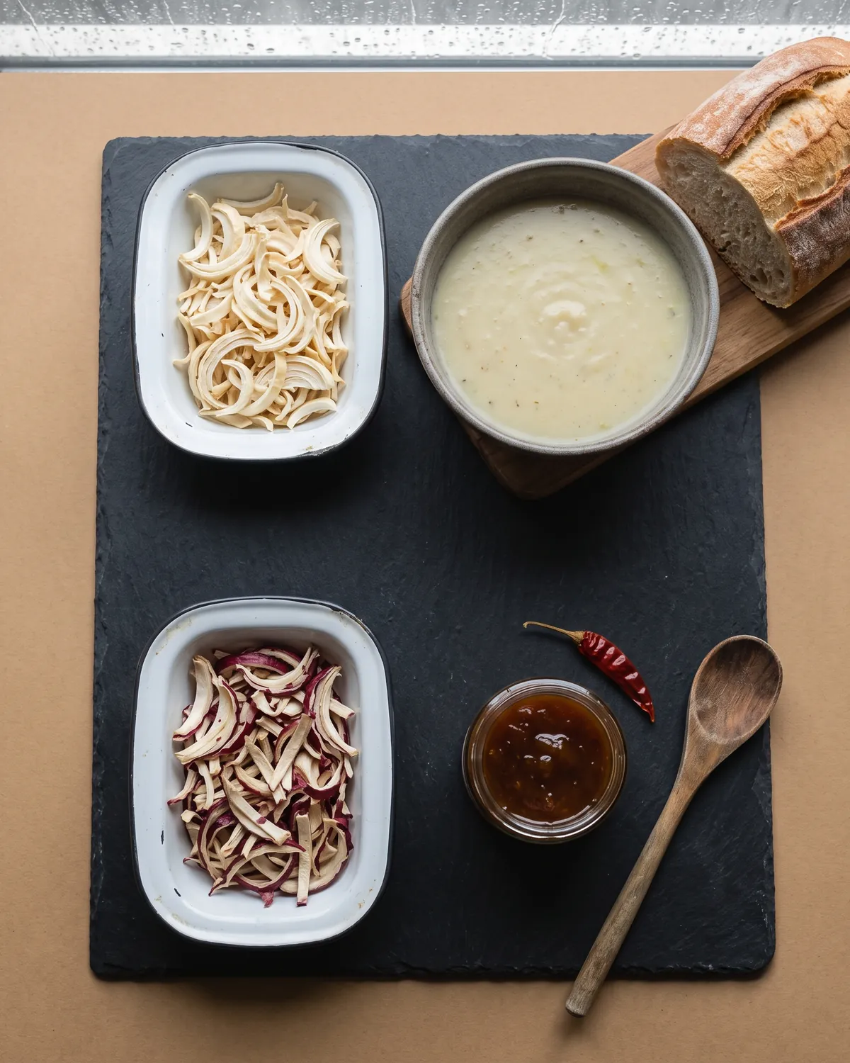 White onion flakes beside a bowl of pale soup and red onion flakes beside an open jar of chutney on a slate board, seen from above.